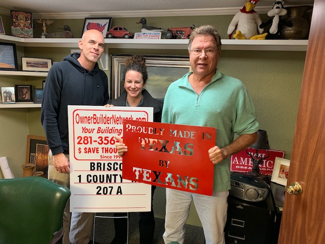 Three people on a man and a woman holding an OwnerBuilderNetwork sign, while and older man holds a 'Proudly made in Texas by texans' sign next to them standing