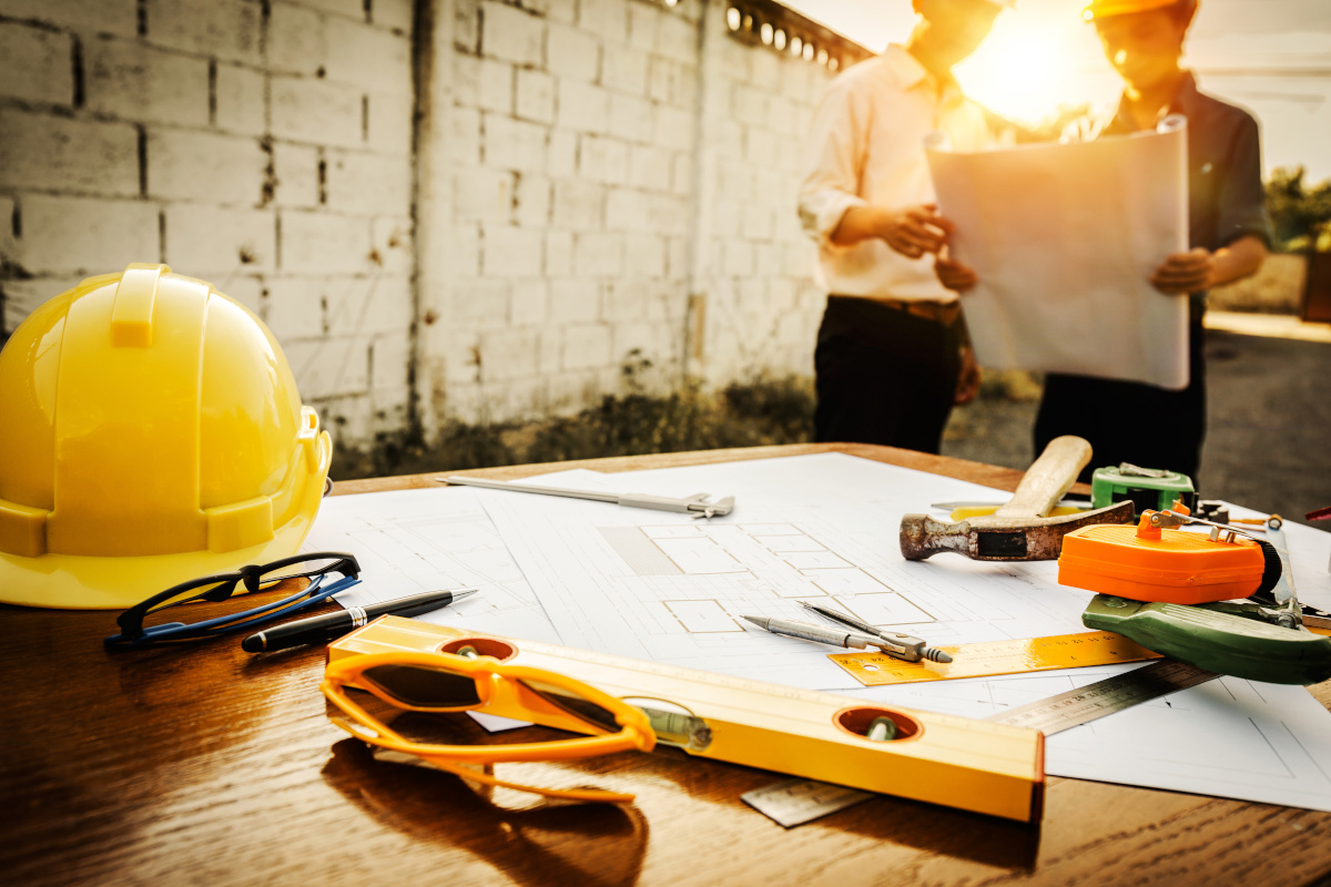 A close up of a table covered with construction gear, with two workers in the background looking over plans