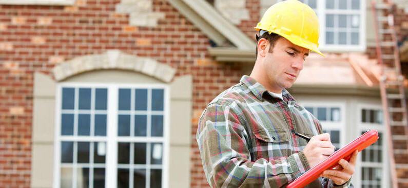 Contractor in Yellow Hard Hat Writing on Red Clipboard in Front of Brick House