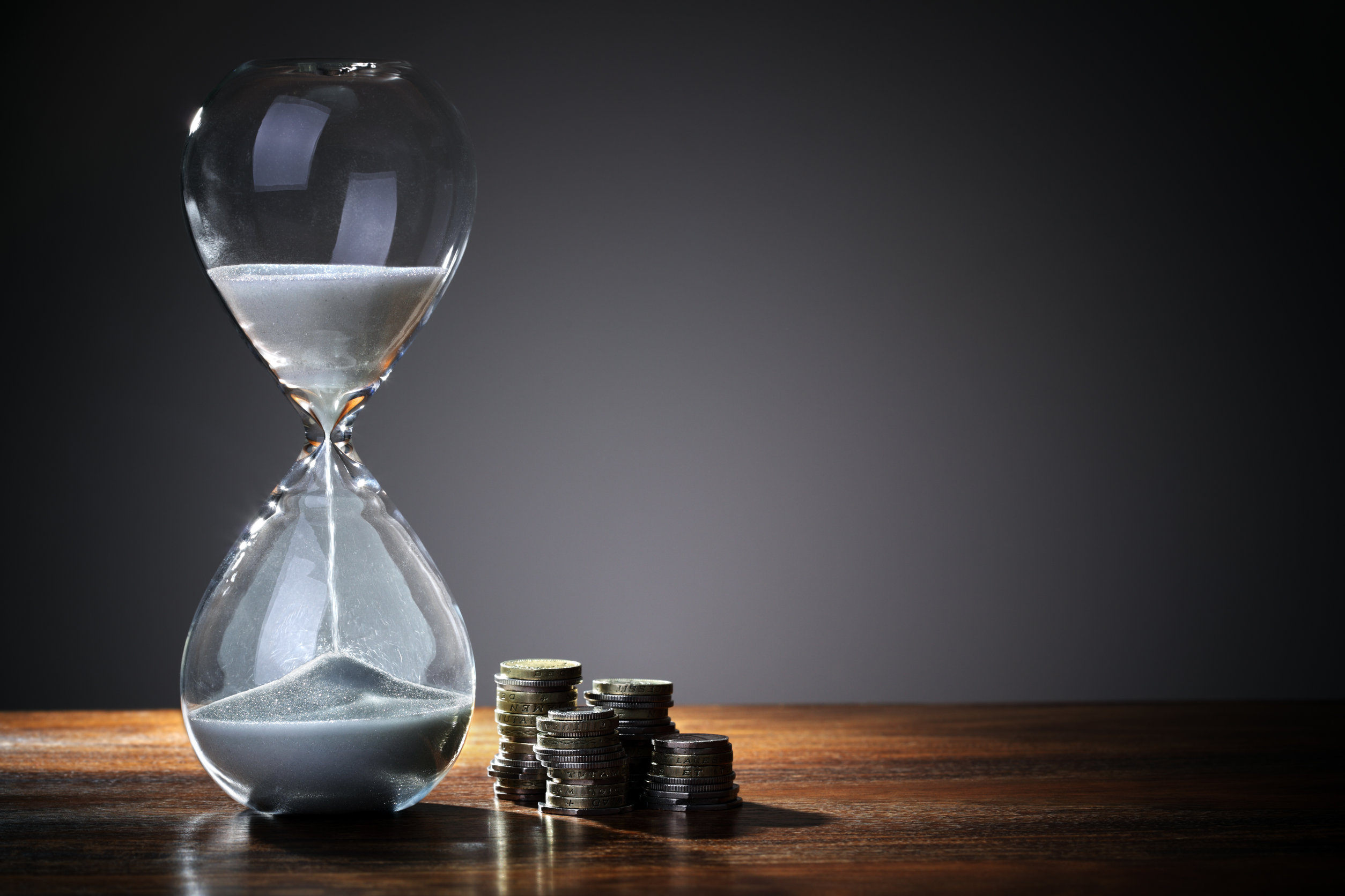 Hourglass on wooden table next to a stack of coins